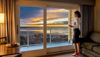 A woman stands in a room by a large window, overlooking a scenic beach sunset, holding a drink, with a meal on the table beside her.