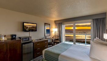 A cozy hotel room with a bed, dresser, TV, mini-fridge, and a large window showcasing a stunning ocean sunset view.