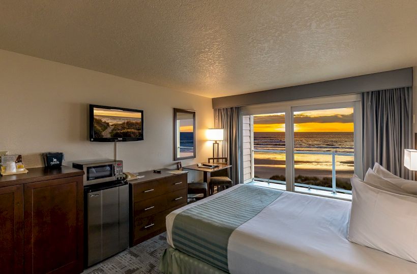 A cozy hotel room with a bed, dresser, TV, mini-fridge, and a large window showcasing a stunning ocean sunset view.