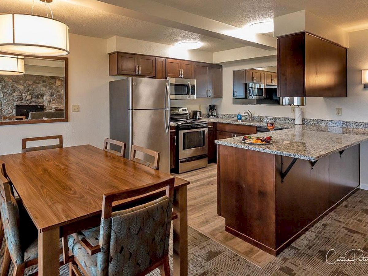 The image shows a modern kitchen and dining area with stainless steel appliances, wooden furniture, and a granite countertop, under warm lighting.