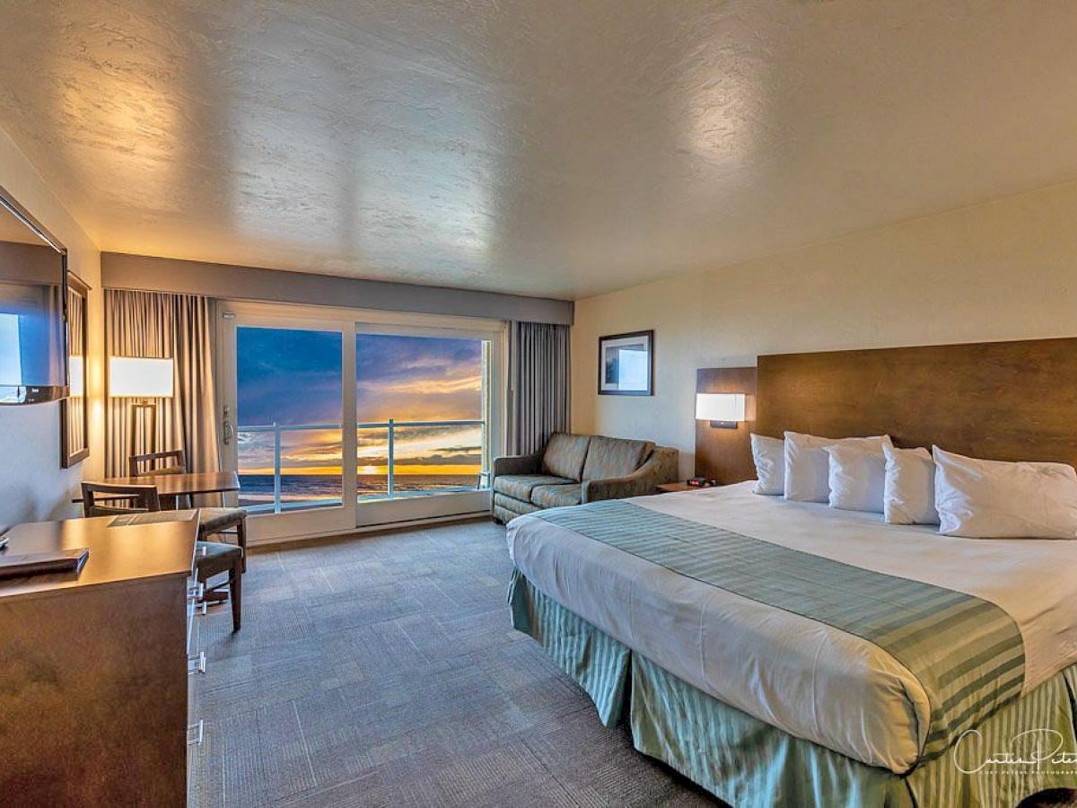 A hotel room features a king-sized bed, a sofa, a TV, a desk, and a sliding door with an ocean view during sunset.