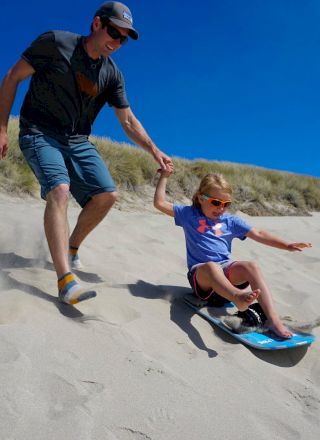 An adult and a child are sandboarding down a sandy hill, with the adult holding the child's hand for support.