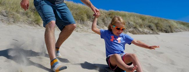 An adult and a child are sandboarding down a sandy hill, with the adult holding the child's hand for support.