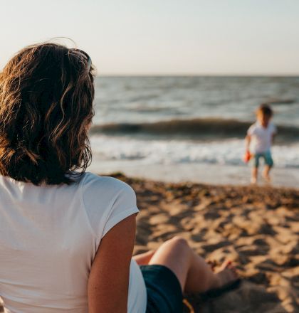 A person with short hair sits on a sandy beach, watching a child play near the water. The ocean waves add a sense of calm to the scene.