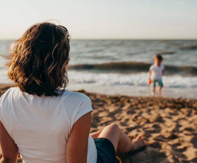 A person with short hair sits on a sandy beach, watching a child play near the water. The ocean waves add a sense of calm to the scene.