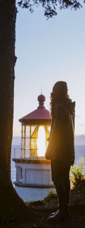 A silhouette of a person stands near a tree overlooking a lighthouse by the sea during sunset, bathed in the warm light of the setting sun.