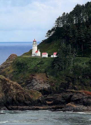 A lighthouse perched on a rocky cliff, surrounded by ocean waves and bordered by a forested area under a cloudy sky.