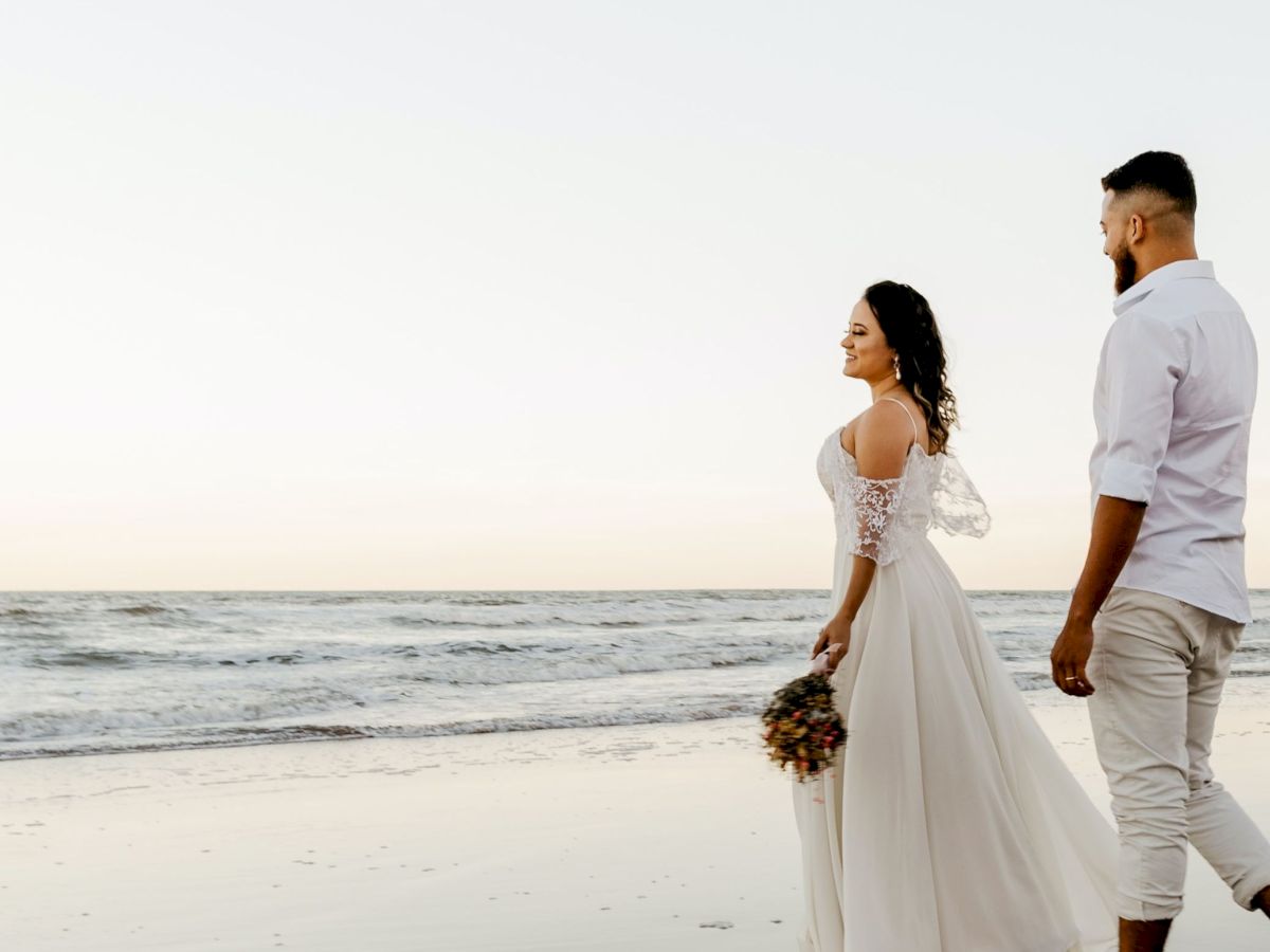 A couple walks along a beach, with the woman in a white dress and the man in light clothing, holding hands during sunset.
