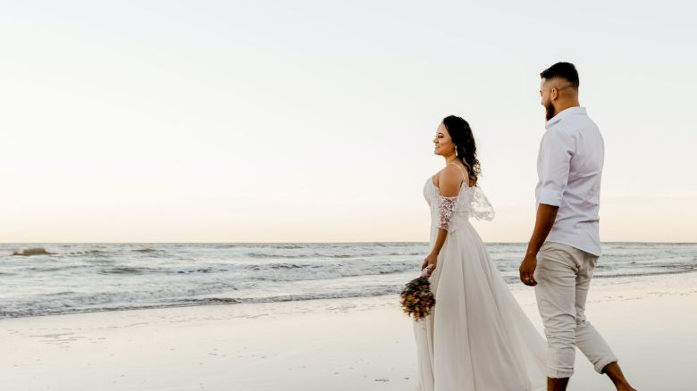 A couple walks along a beach, with the woman in a white dress and the man in light clothing, holding hands during sunset.