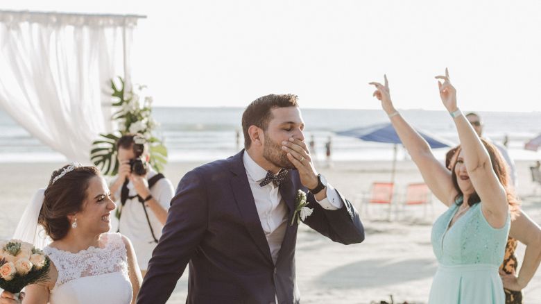 A bride and groom hold hands at a beach wedding, with bridesmaids cheering. The ocean and wedding decor are visible in the background.