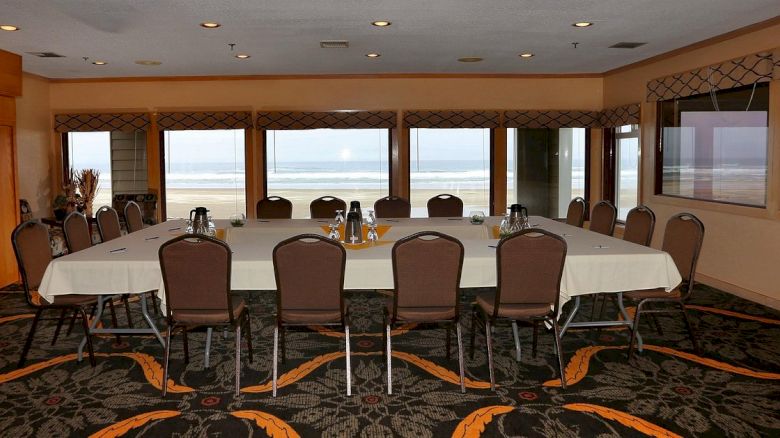 A conference room with a large table, chairs, and refreshments, overlooking a beach through windows, creating a meeting space with an ocean view.