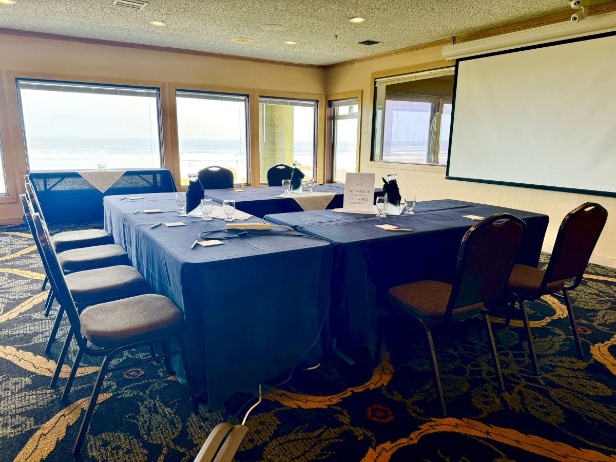A conference room setup with tables in a U-shape, chairs, a screen, and a window view of the outside scenery.
