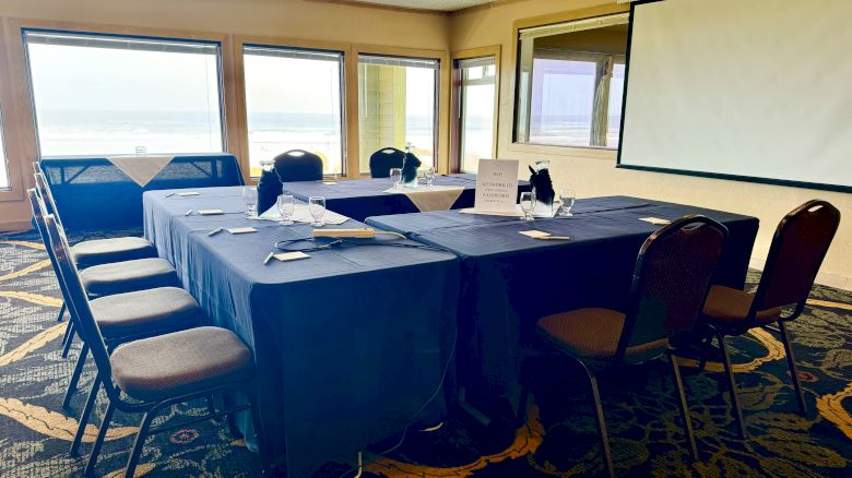 A conference room setup with tables in a U-shape, chairs, a screen, and a window view of the outside scenery.