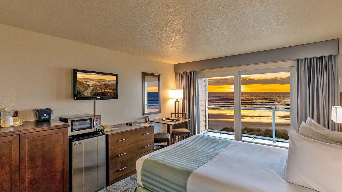 A cozy hotel room with a bed, a TV, a desk, and oceanfront windows displaying a sunset.
