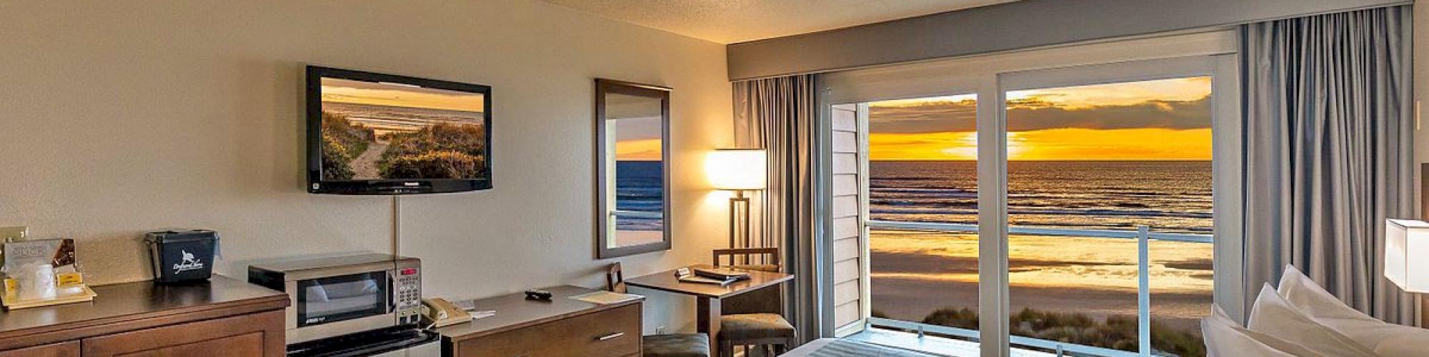 A cozy hotel room with a bed, a TV, a desk, and oceanfront windows displaying a sunset.