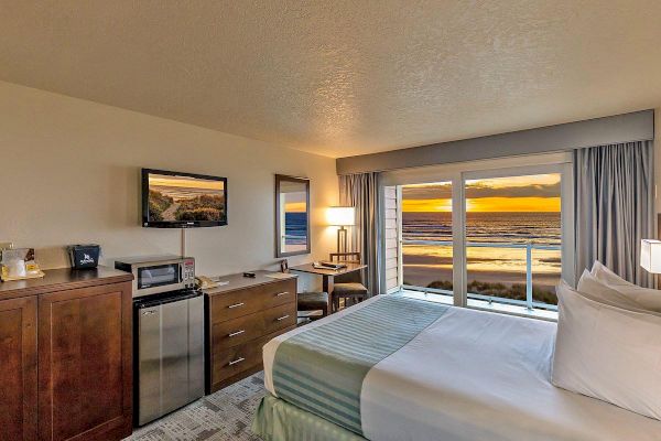 The image shows a cozy hotel room with a bed, TV, desk, mini fridge, and a stunning sunset view over an ocean through large windows.