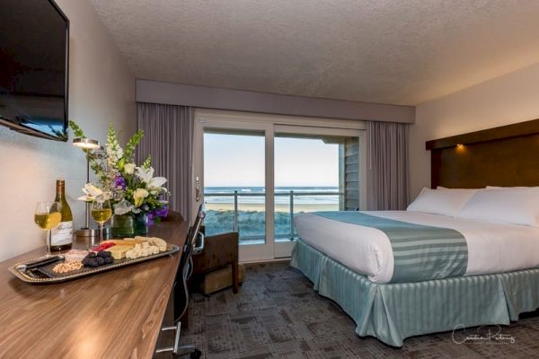 A hotel room with a king-sized bed, a balcony overlooking the ocean, a desk with flowers, wine, and cheese, and a wall-mounted TV always ending the sentence.