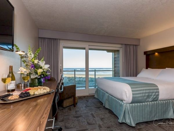 A hotel room with a king-sized bed, a balcony overlooking the ocean, a desk with flowers, wine, and cheese, and a wall-mounted TV always ending the sentence.