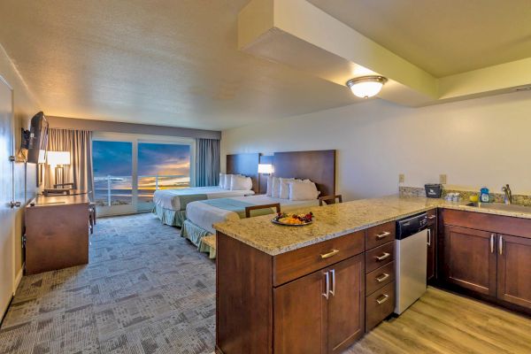 A spacious hotel room features two beds, a kitchenette with granite countertops, a small dining area, and a scenic sunset view through large windows.