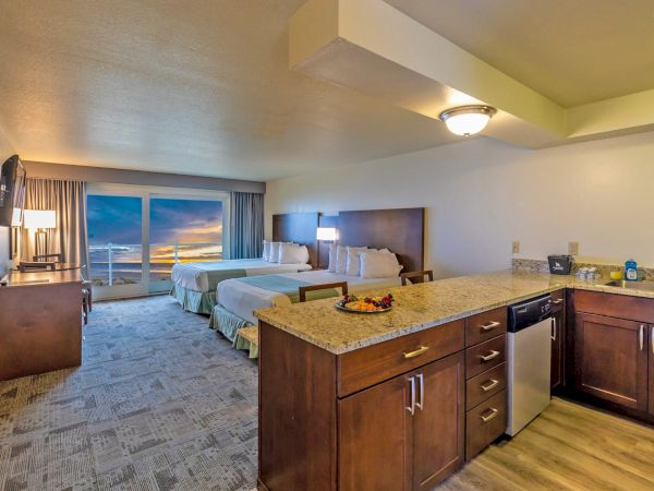 A spacious hotel room features two beds, a kitchenette with granite countertops, a small dining area, and a scenic sunset view through large windows.
