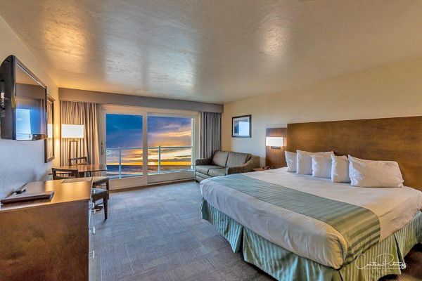 A hotel room with a king-sized bed, a couch, a TV, a desk, and a large window displaying a sunset view over the ocean.