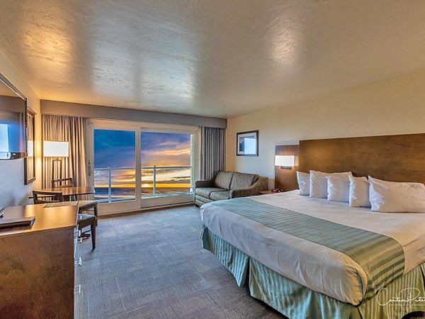 A hotel room with a king-sized bed, a couch, a TV, a desk, and a large window displaying a sunset view over the ocean.
