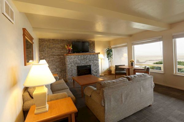 A cozy living room with a stone fireplace, two sofas, lamps, a TV, large windows with a scenic view, and a wooden table with chairs.