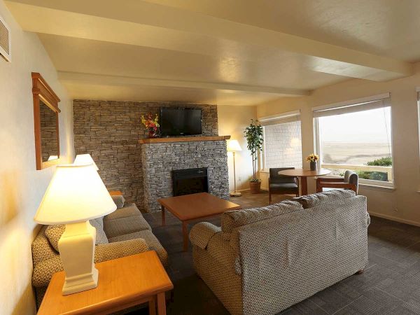 A cozy living room with a stone fireplace, two sofas, lamps, a TV, large windows with a scenic view, and a wooden table with chairs.