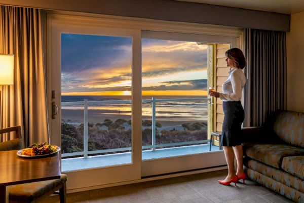 A woman stands by a sliding glass door in a hotel room with a beach view at sunset, holding a drink. A table with food is nearby.