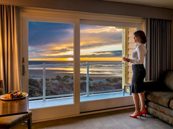 A woman stands by a sliding glass door in a hotel room with a beach view at sunset, holding a drink. A table with food is nearby.