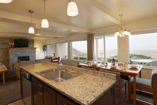 This image shows a spacious kitchen and dining area with a granite island, large windows with an ocean view, and modern furnishings.