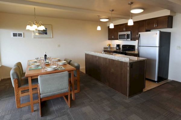 This image depicts a modern kitchen and dining area with a wooden table, gray chairs, and a kitchen island with stainless steel appliances.