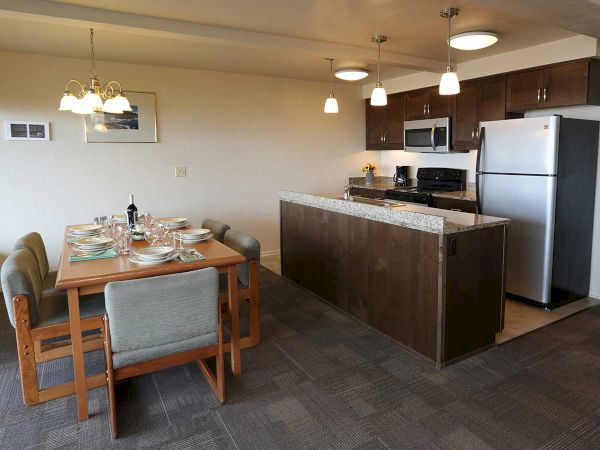 This image depicts a modern kitchen and dining area with a wooden table, gray chairs, and a kitchen island with stainless steel appliances.