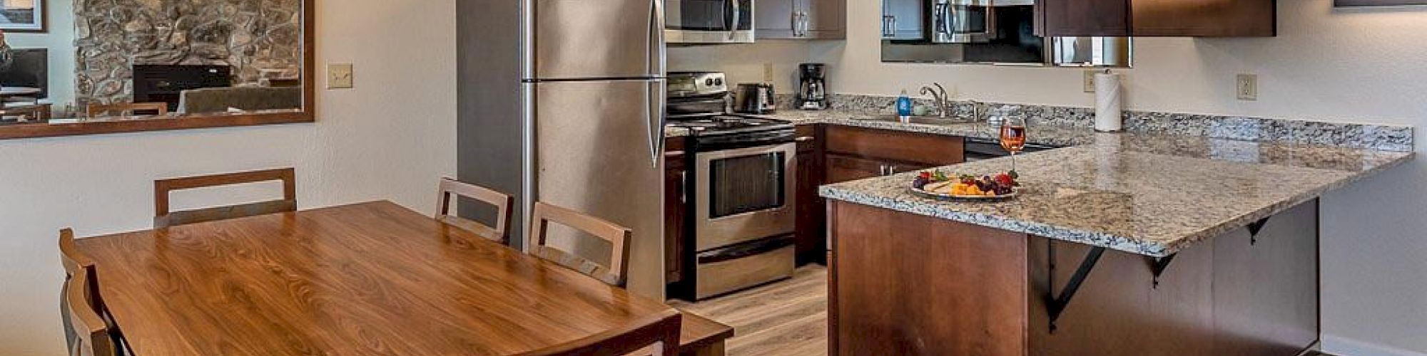 The image shows a modern kitchen and dining area with wooden cabinets, stainless steel appliances, a large dining table, and overhead lighting.