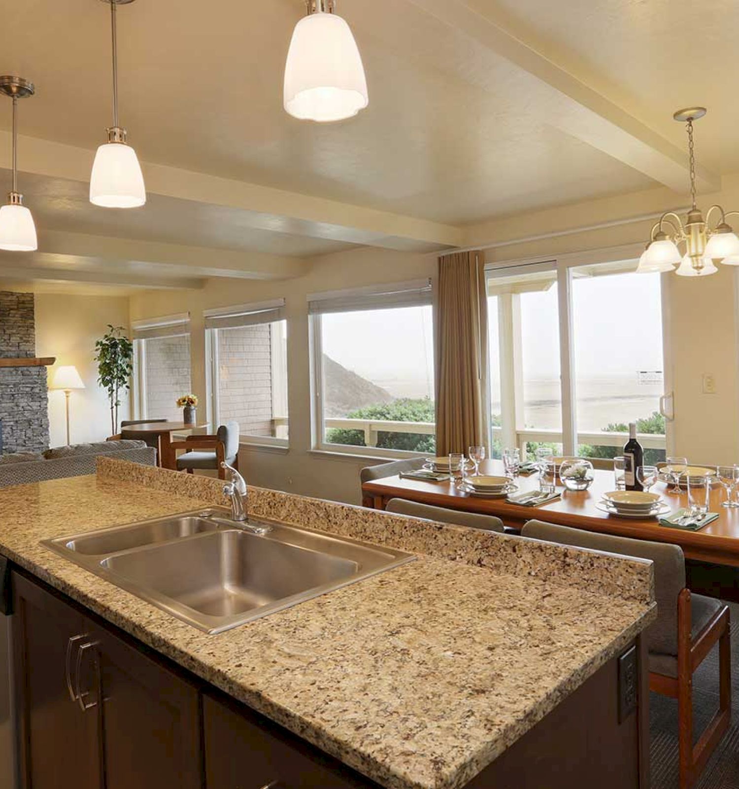 A bright living-dining room with large windows, a stone fireplace, an island kitchen with granite countertops, and a set dining table.