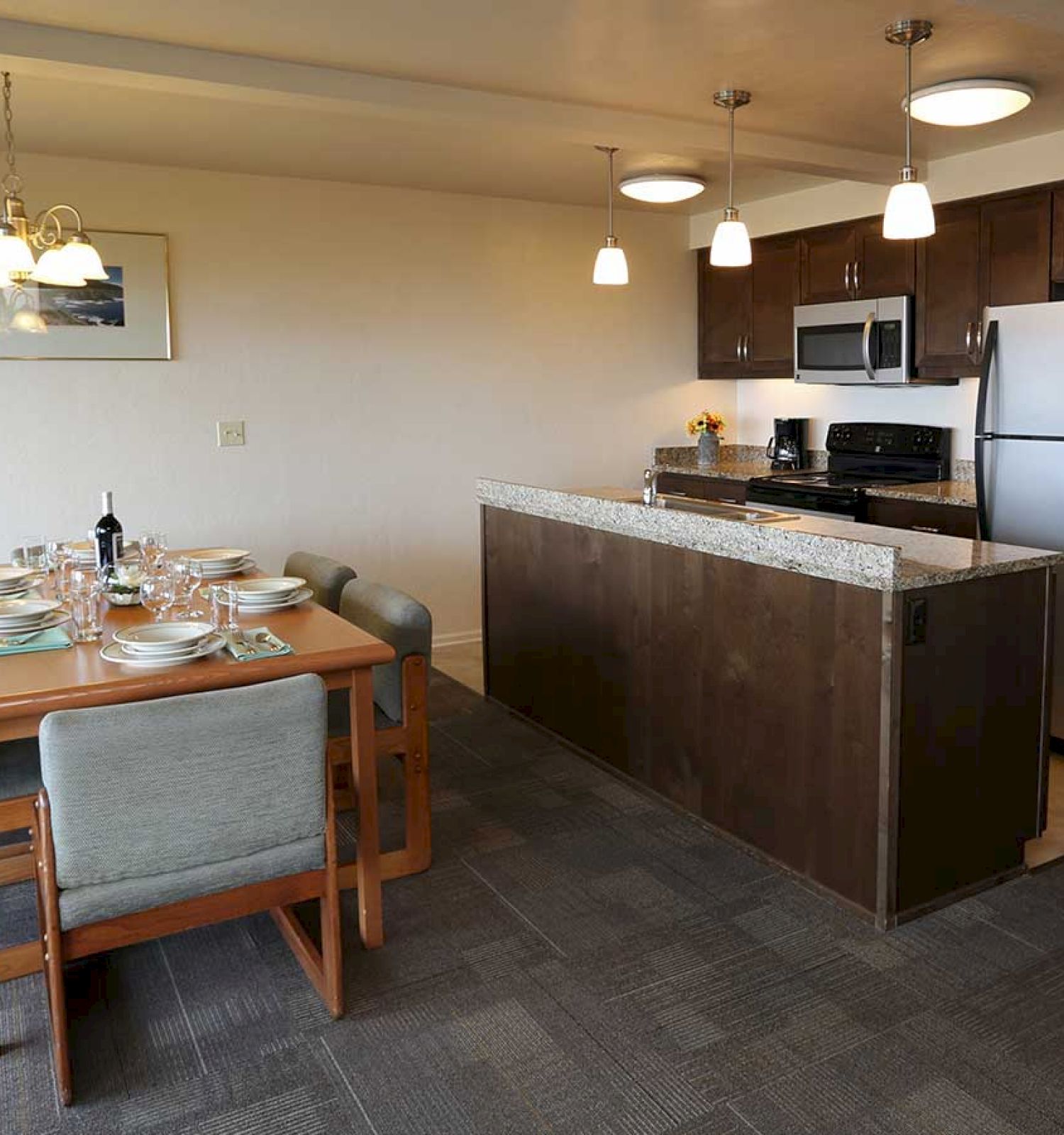 A modern kitchen and dining area with a wooden table set for six, a stainless steel fridge, and overhead lighting in a spacious, carpeted room.