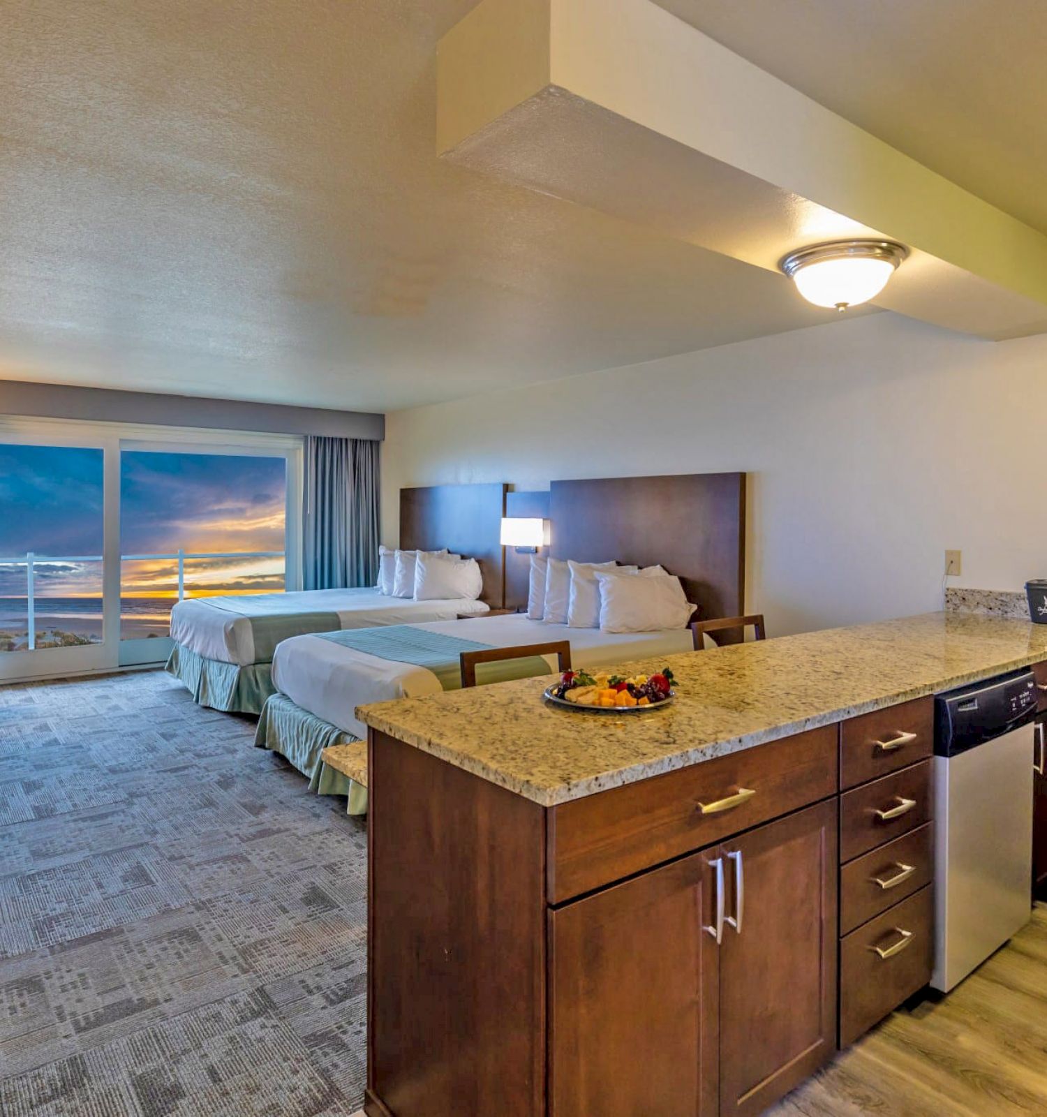 A hotel room with two beds, a kitchenette, and a view of the sunset through large windows, featuring modern amenities and a comfortable interior.