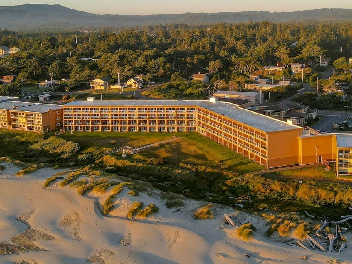 The image shows an aerial view of a beachfront hotel with multiple stories, surrounded by sand dunes, greenery, and residential buildings in the distance.