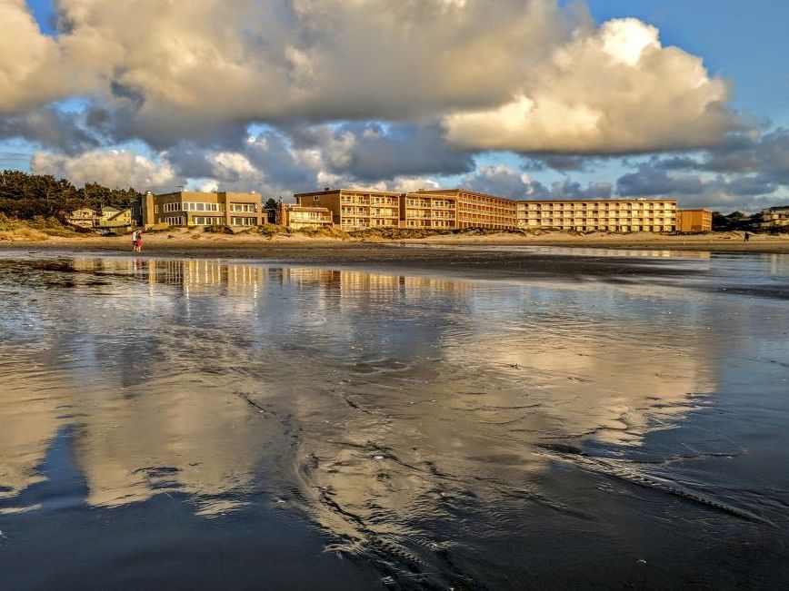 A beach scene with buildings in the background and a cloudy sky reflected in the water, creating a serene, picturesque view.