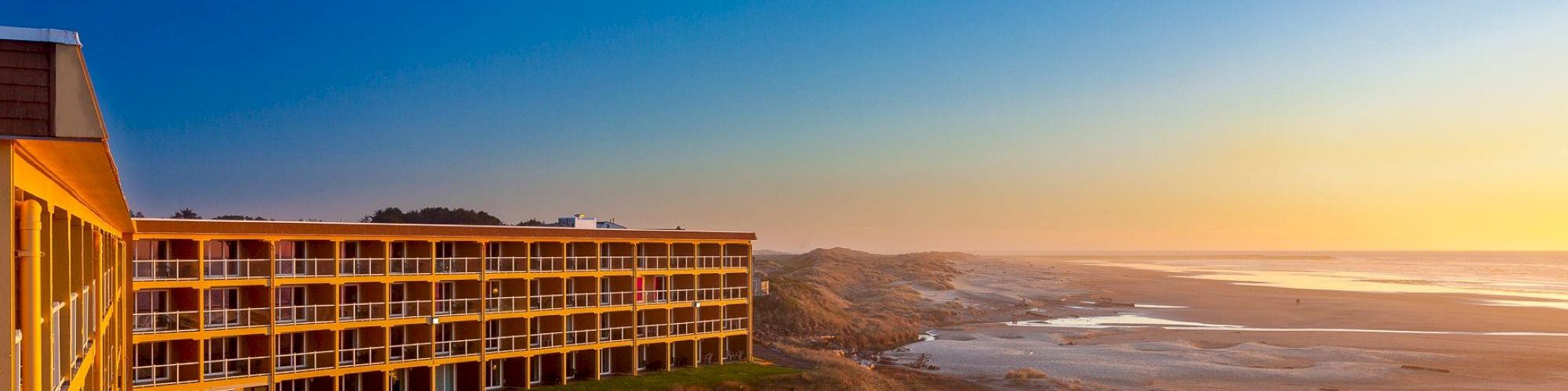 A coastal resort building near a sandy beach with grassy dunes, overlooking a serene ocean view during a colorful sunset.