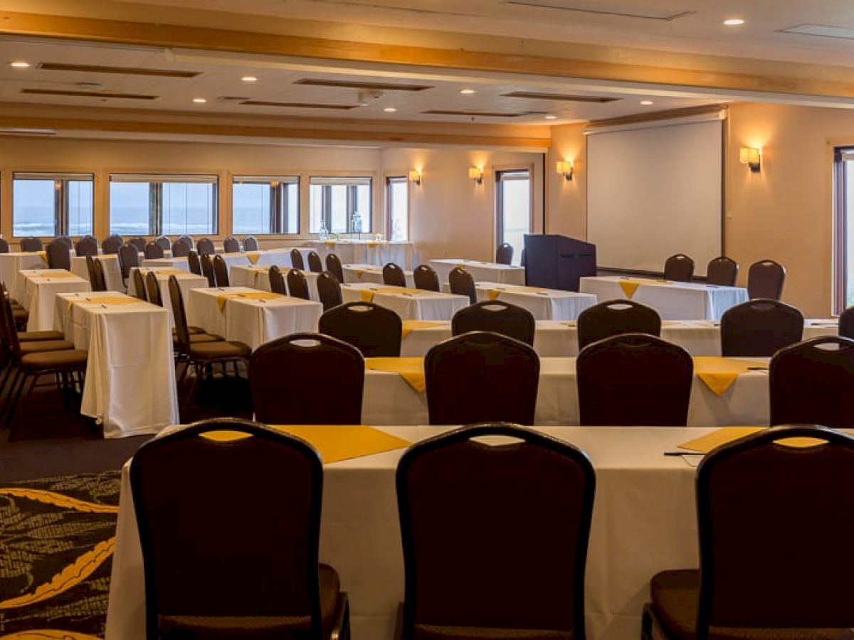 A conference room is set up with rows of tables and chairs facing a podium and projection screen, with tablecloths and yellow accents.