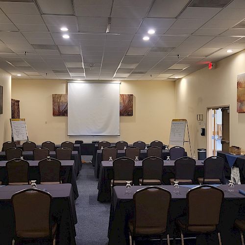 A conference room with rows of chairs and tables facing a projection screen, flip charts, and a small table on the side with a lamp.