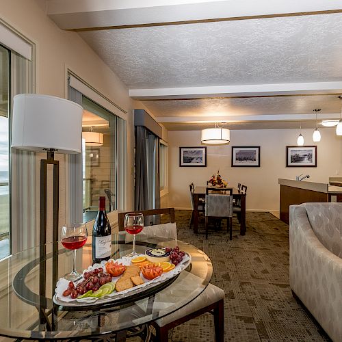 An open-plan living area with a coastal view, a dining set, a kitchen, and a table with wine, grapes, and cheese.