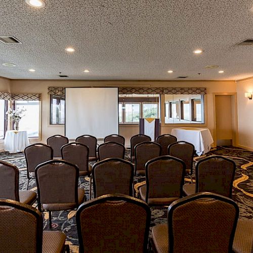 The image shows a small conference room setup with rows of chairs facing a projector screen, large windows, and a table in the back.