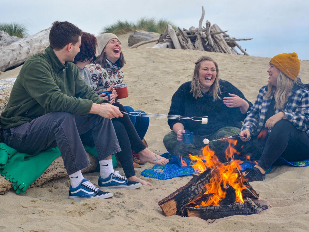 A group of people sitting around a campfire on a beach, laughing and roasting marshmallows.