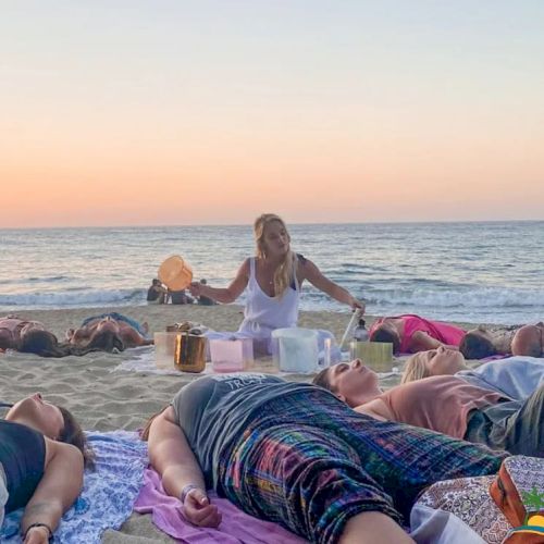 People are lying on the beach during sunset while a person in the center performs a ritual or meditation session with bowls.