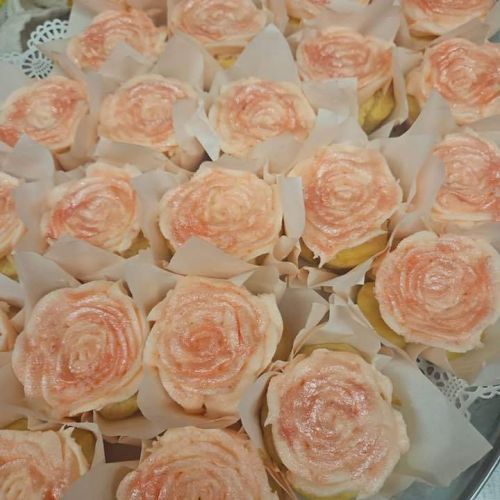 The image shows a tray of cupcakes with pink rose-shaped frosting on top, each placed in white wrappers, arranged closely together.