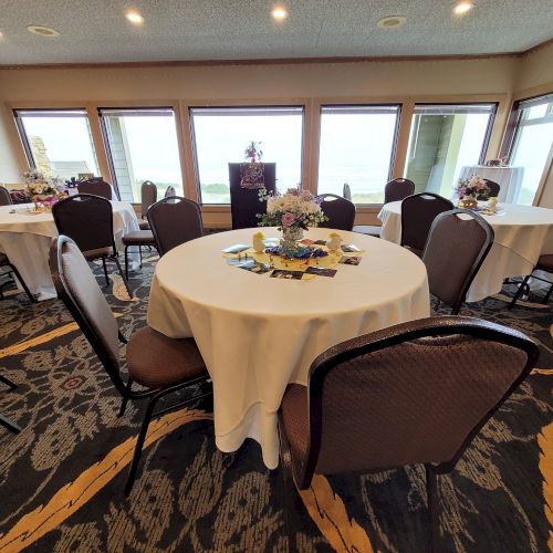 A small event room with round tables covered in white tablecloths, chairs, floral centerpieces, and table settings, set against large windows with outside views.