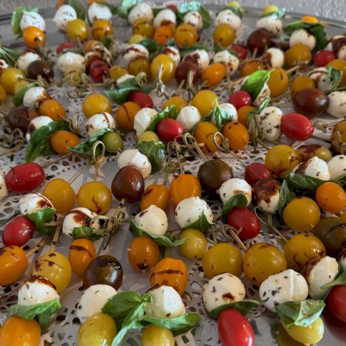 The image shows a tray of skewers with cherry tomatoes, mozzarella balls, basil leaves, and a balsamic drizzle.