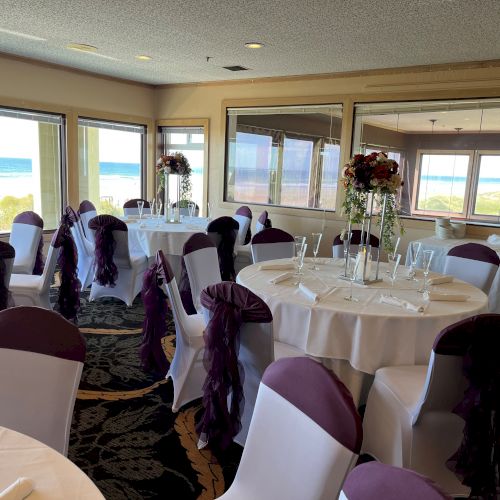 A beautifully decorated dining room with round tables, white tablecloths, and purple chair sashes overlooking a scenic ocean view is shown.